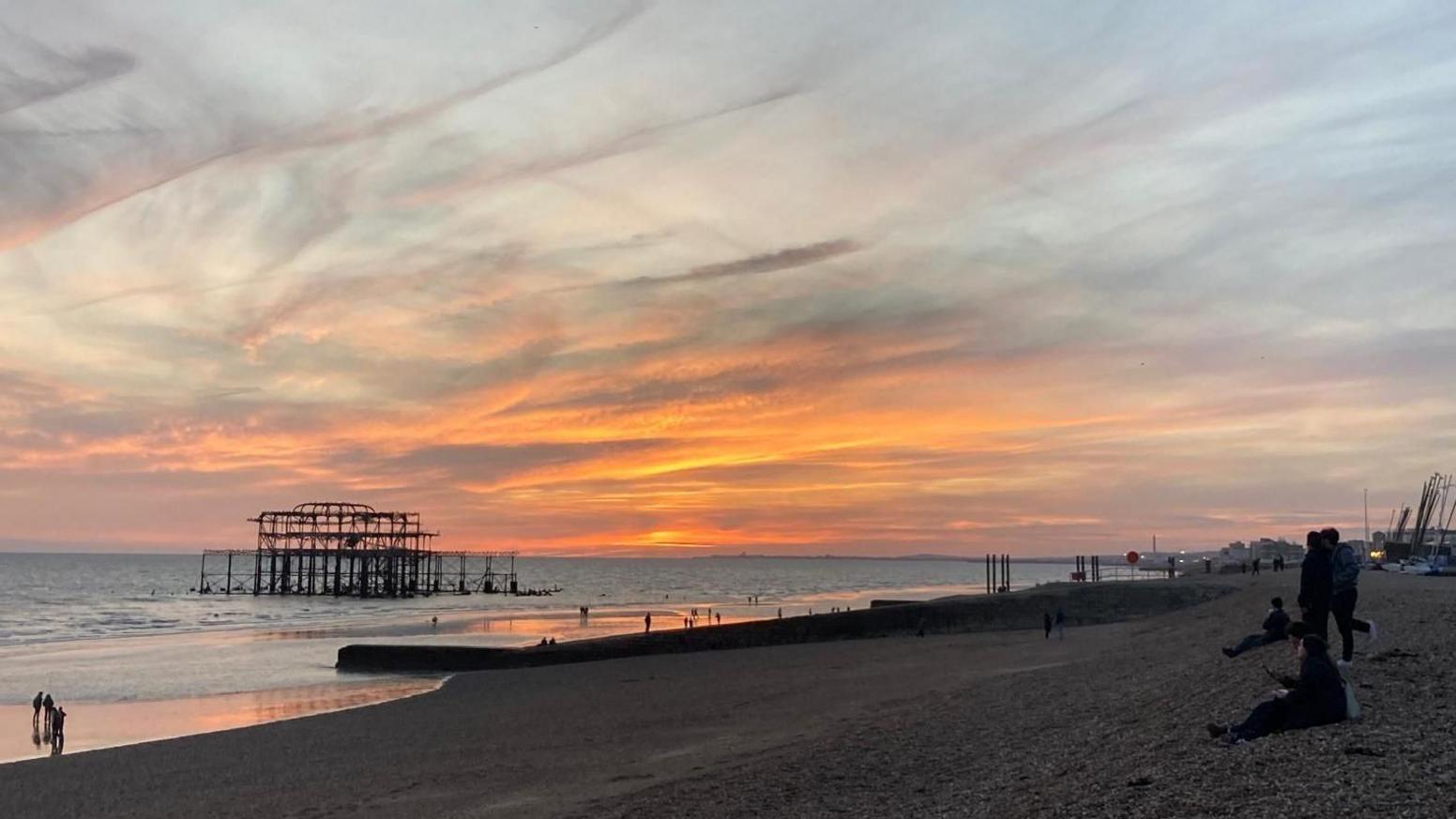 Një fotografi e detit dhe plazhit në perëndim të diellit me shtresa resh të larta në qiell, disa njerëz të ulur në plazh dhe rrënojat e djegura të West Pier të Brighton-it, të dukshme.