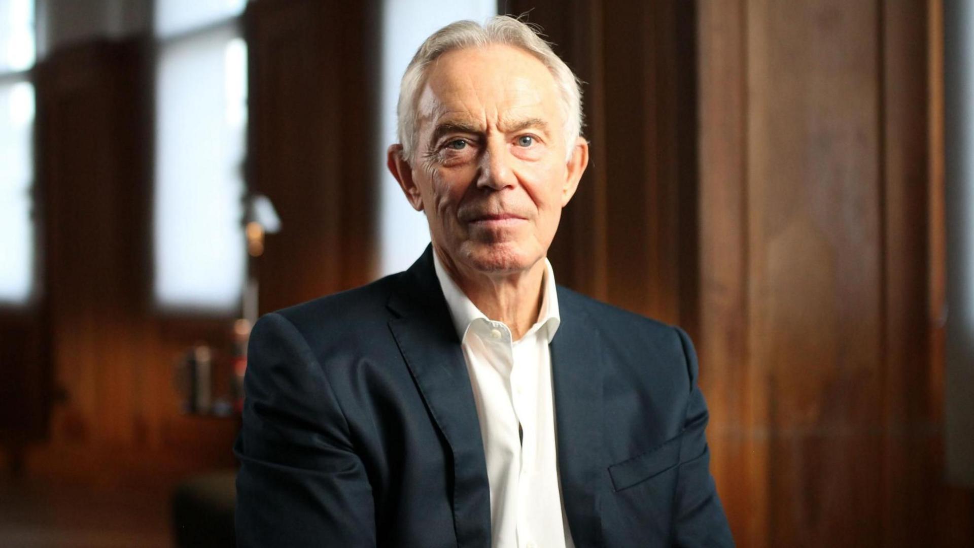 Tony Blair shikon nga kamera. Ai ka veshur një kostum blu marine dhe një këmishë të bardhë me jakë të hapur.
