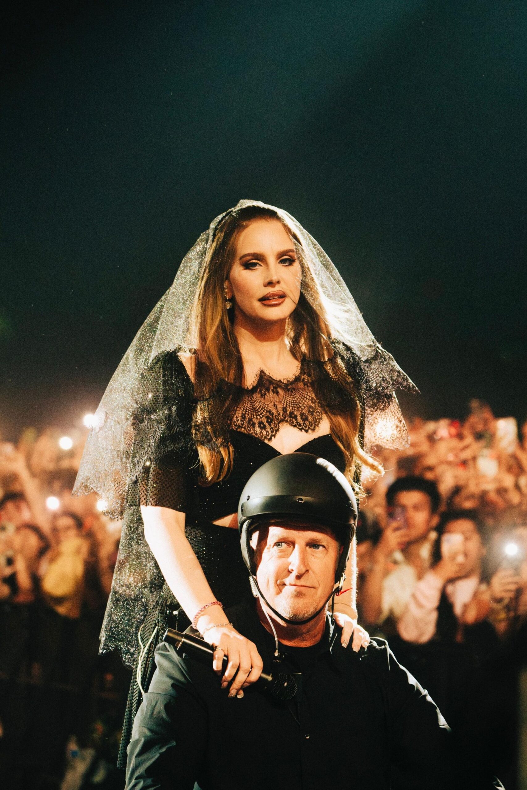 Lana Del Ray, e veshur me rroba të zeza nusërie, qëndron mbi supet e një burri me helmetë dhe xhaketë të zezë, ndërsa turma i fotografon dhe i filmon me telefona celularë.