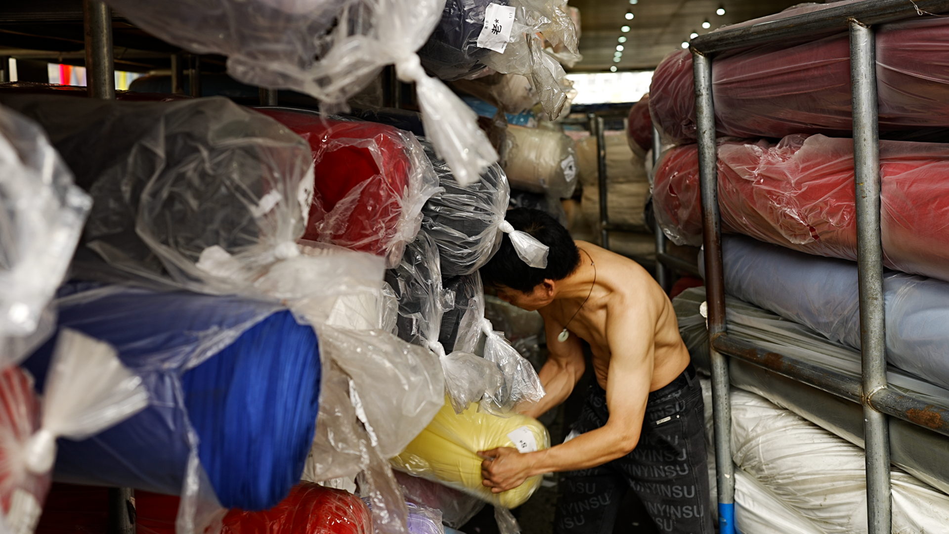 Një burrë pa këmishë me pantallona të zeza përdredh rrotulla të mëdha pëlhure të mbështjella me plastikë, në një depo tekstili në Guangzhou.