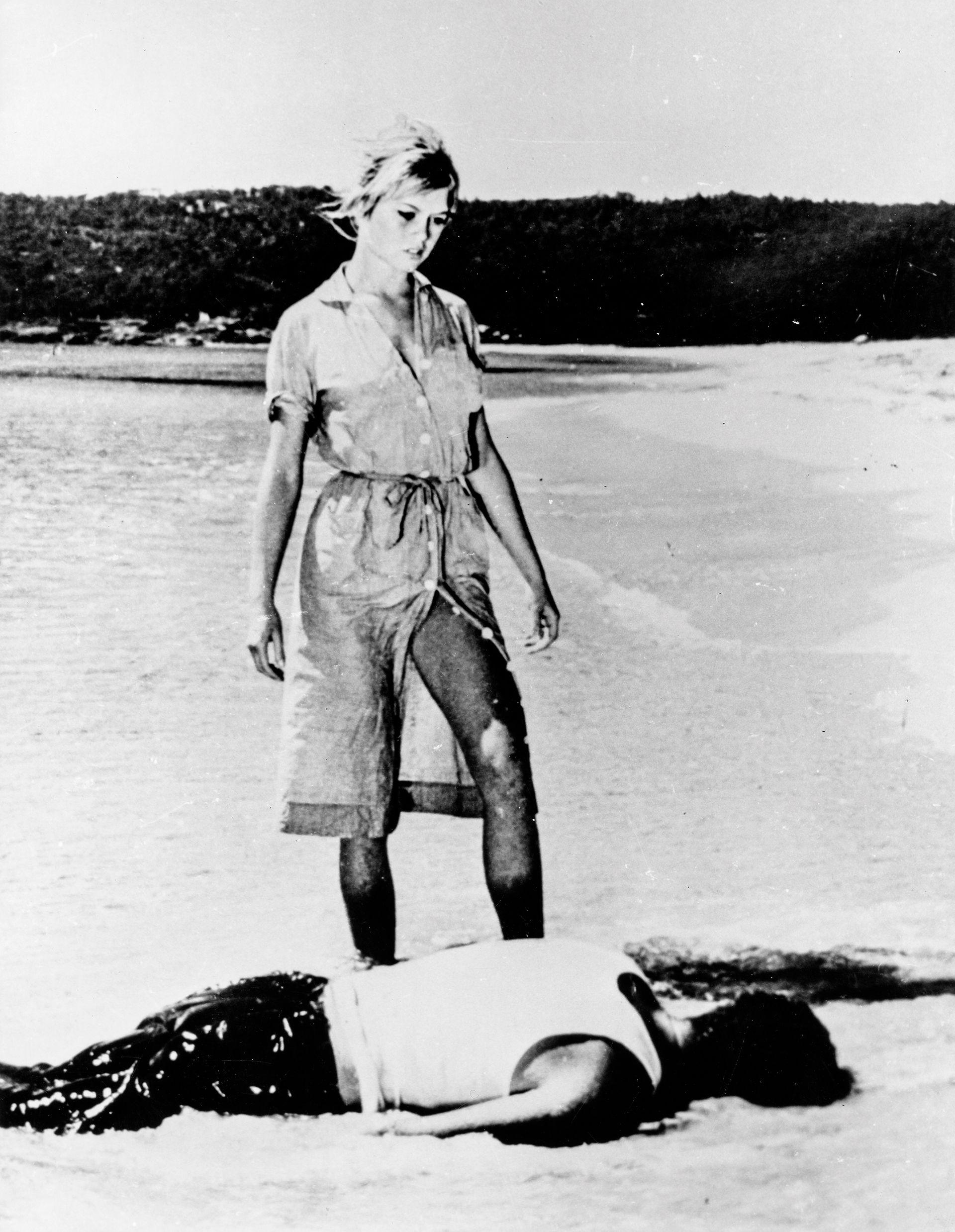 Brigitte Bardot në një pamje nga filmi “Dhe Zoti Krijoi Gruan”, e veshur me një fustan pjesërisht të zbërthyer nga fundi në mënyrë që të hapet për të zbuluar kofshën e saj, ndërsa qëndron mbi një burrë të shtrirë me fytyrë poshtë në plazh.