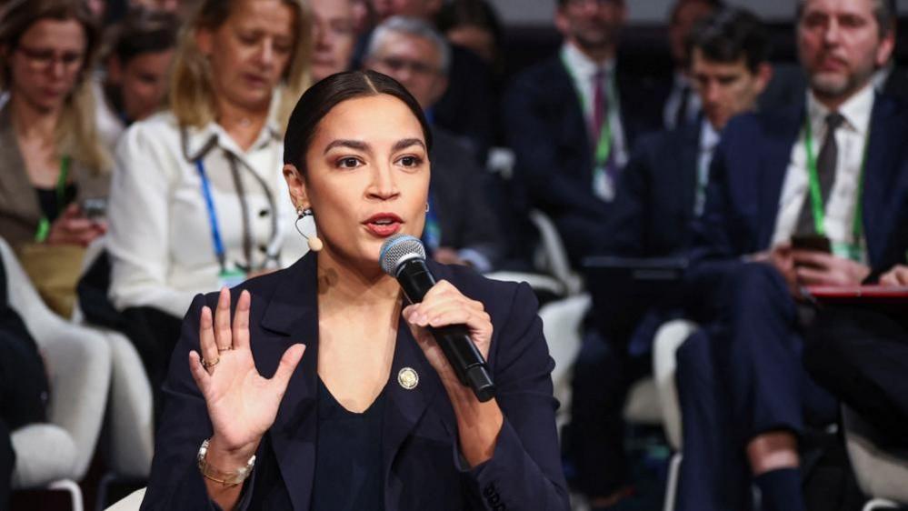 Përfaqësuesja Alexandria Ocasio-Cortez, e veshur me një kostum të errët dhe me flokët e lidhura prapa, mban një mikrofon në njërën dorë dhe bën gjeste me pëllëmbën e hapur me tjetrën përpara një turme njerëzish të ulur.