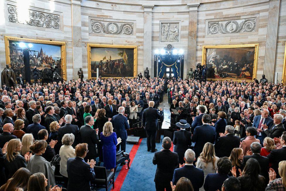 Trump qëndron në një podium në Rotondë, i cili është plot me të ftuar. Ai na ka kthyer shpinën dhe duket sikur po bën një gjest festiv me grushtin e djathtë lart.
