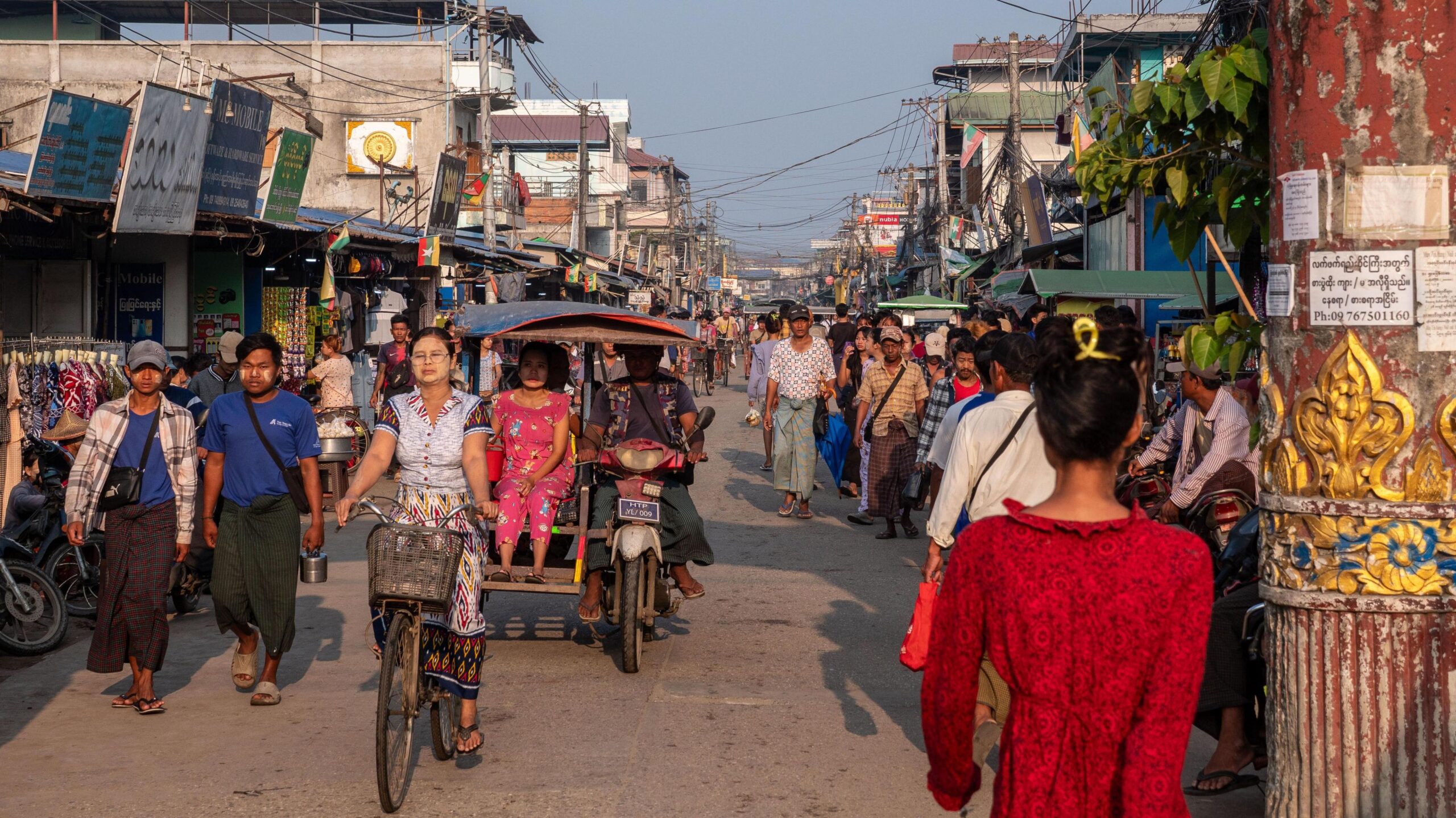 Një rrugë e ngarkuar në një treg në Yangon. Njerëzit shihen duke ecur përgjatë saj, pranë rikshave që transportojnë njerëz.