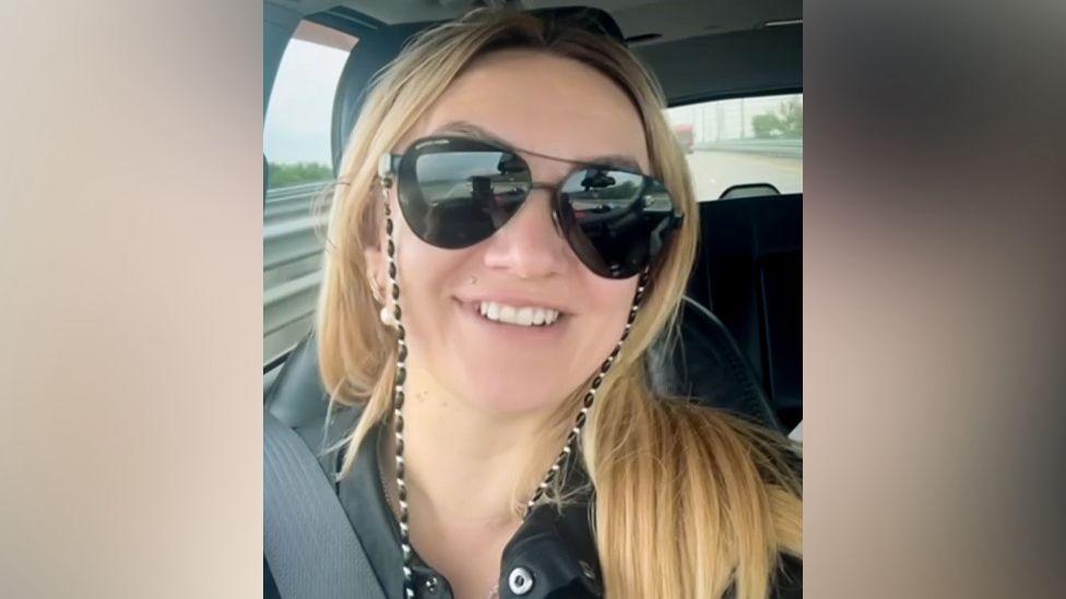 Polina Azarnykh duke shikuar drejt e në kamerë ndërsa është ulur në një automjet. Ajo ka flokë të gjatë bjondë dhe mban syze dielli të mëdha, një bluzë të zezë dhe është duke buzëqeshur.