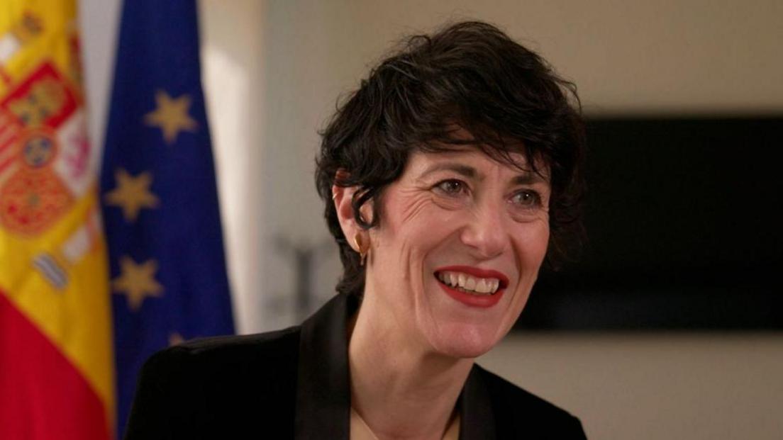 Elma Saiz, Ministrja për Përfshirjen, Sigurimet Shoqërore dhe Imigracionin, buzëqesh para flamujve spanjollë dhe të BE-së