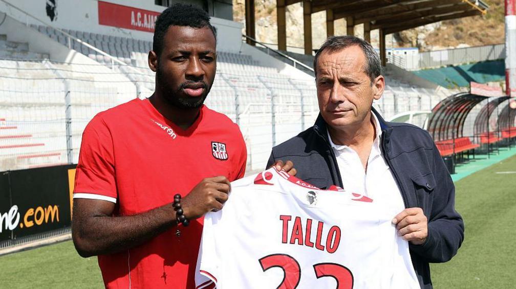 Një futbollist i Bregut të Fildishtë me një bluzë të kuqe futbolli qëndron me një burrë më të vjetër në të djathtë, ndërsa të dy mbajnë një bluzë të bardhë me etiketën Tallo 22