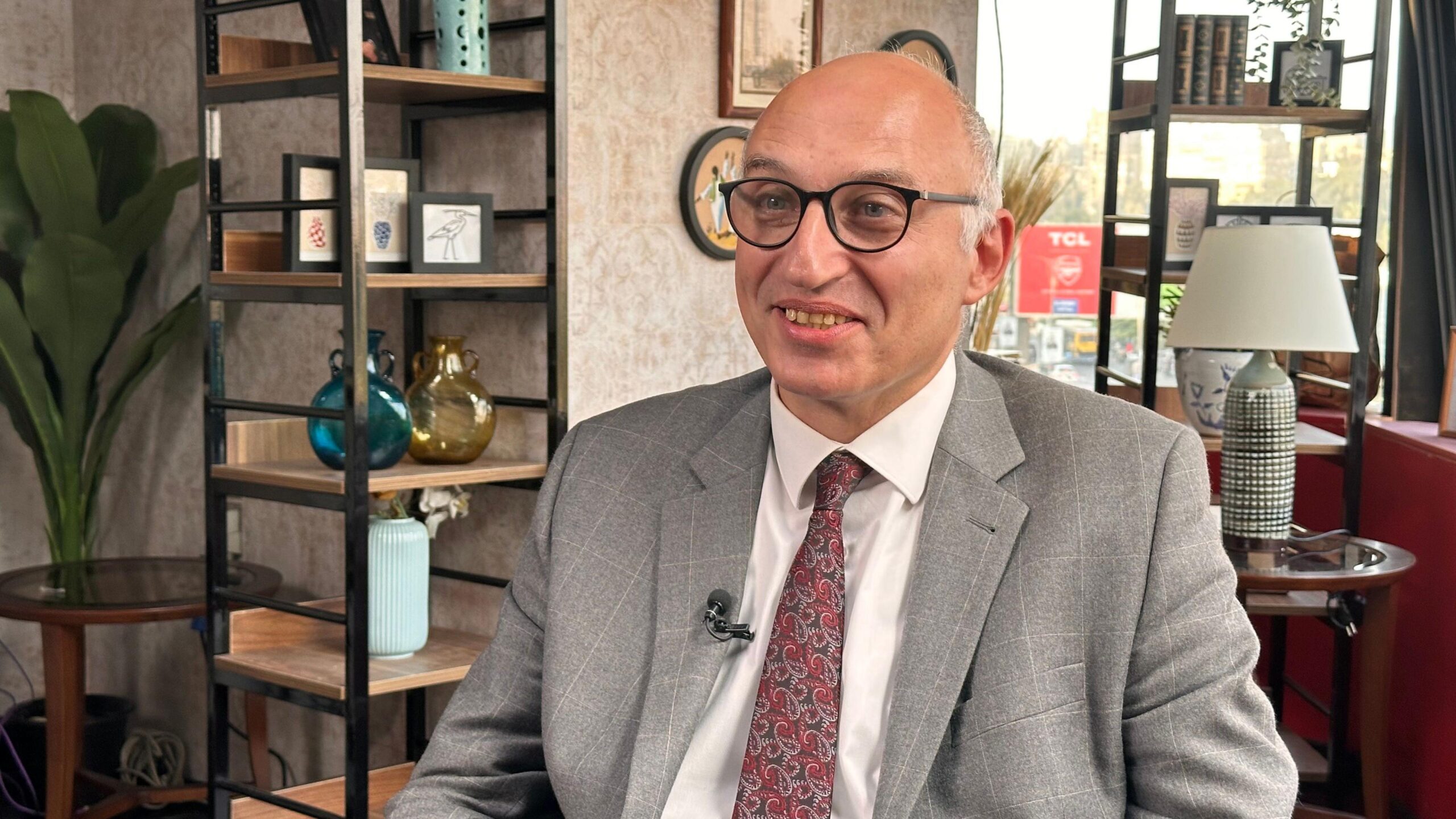 Dr. Tarek Tawfik mban veshur një kostum gri, kravatë të kuqe dhe syze. Ai buzëqesh paksa nga kamera. Pas tij, zyra e tij është e mbushur me rafte të veçantë që mbajnë vazo, libra dhe korniza fotografish.