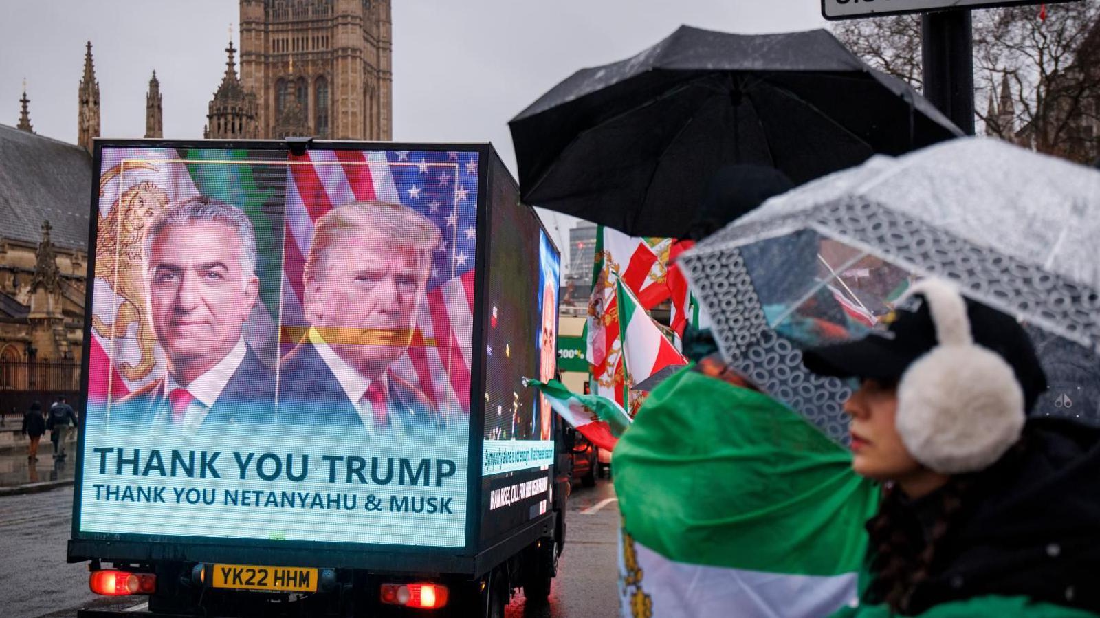 Një billboard i lëvizshëm me mbishkrimin "Faleminderit Trump" kalon pranë mbështetësve të Reza Pahlavi, djalit në mërgim të shahut të fundit të Iranit, i cili proteston jashtë Parlamentit në Londër më 13 janar 2026.