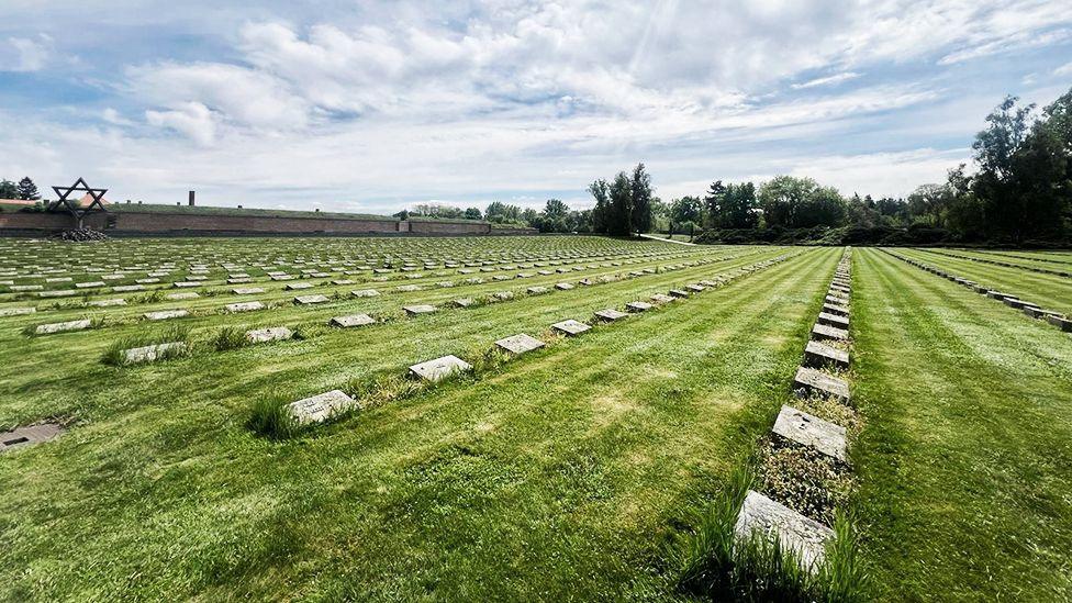 Varreza në Theresienstadt, një ish-kamp përqendrimi nazist, ku shihen rreshta me gurë varresh të sheshtë të rregulluar në vija paralele përgjatë një lëndine të gjelbër. Një monument i madh i Yllit të Davidit qëndron dukshëm në sfond, duke simbolizuar kujtimin hebre. Vendi përkujton viktimat e Holokaustit, me pemë dhe një qiell pjesërisht me re që rrethojnë peizazhin solemn.