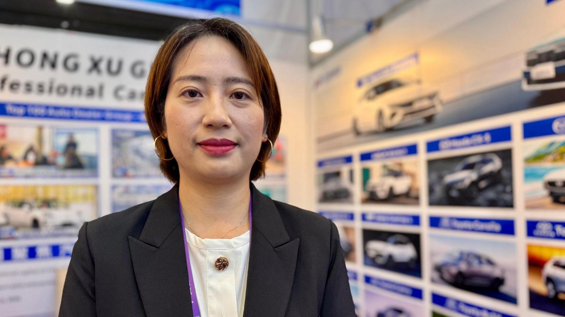 Joyce Liu në një stendë makinash elektrike. Joyce ka flokë të shkurtër ngjyrë kafe dhe mban veshur një këmishë të bardhë dhe një xhaketë të zezë elegante, me fytyrë drejtpërdrejt nga kamera.