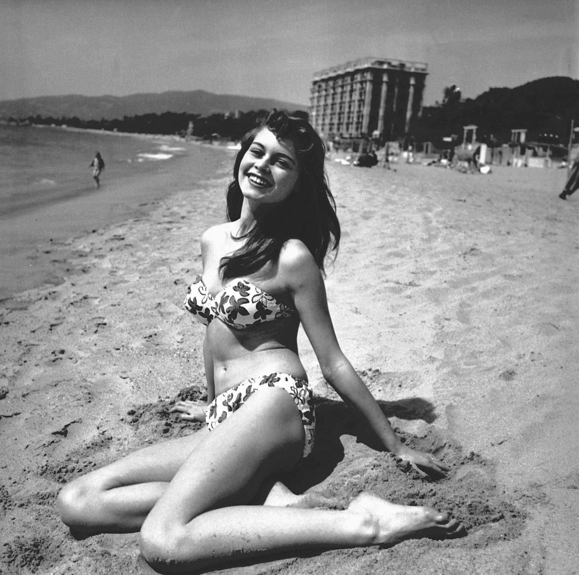 Brigitte Bardot e fotografuar bardh e zi, me flokë të gjatë të errët, e veshur me bikini me lule, ndërsa ulet në rërë duke buzëqeshur për fotografët në Festivalin e Filmit në Kanë në vitin 1953.