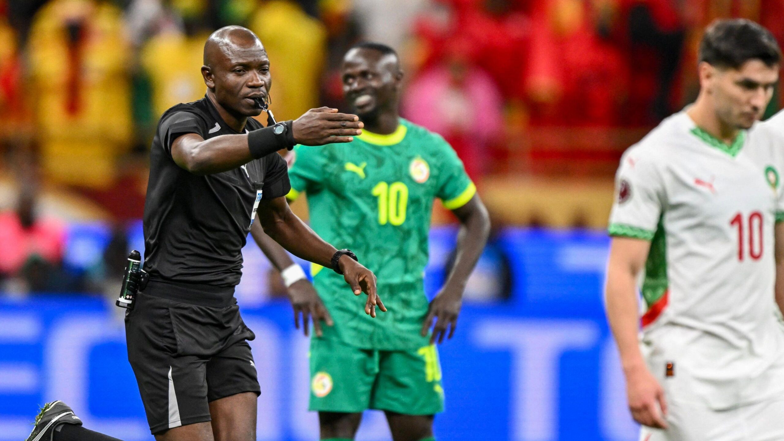 Arbitri Jean-Jacques Ndala drejton krahun e djathtë përpara ndërsa jep një penallti në finalen e Kupës së Kombeve të Afrikës. Jashtë fokusit në sfond mund të shihet Sadio Mane me një buzëqeshje ironike në fytyrë.