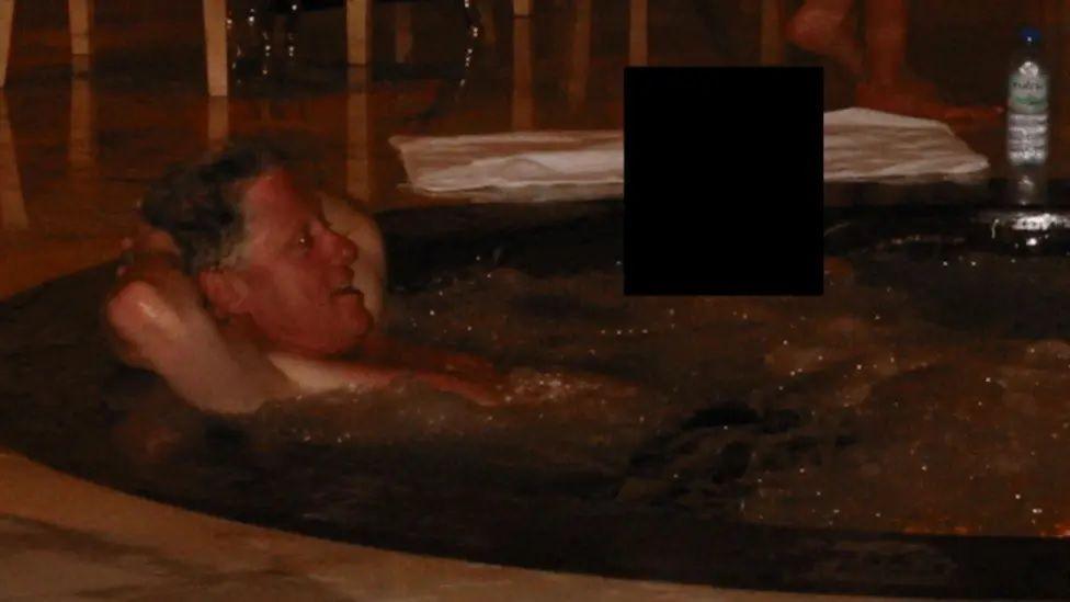 Bill Clinton është ulur në një vaskë të nxehtë me duart pas kokës. Një person tjetër është ulur afër, por fytyra e tij është e nxirë.