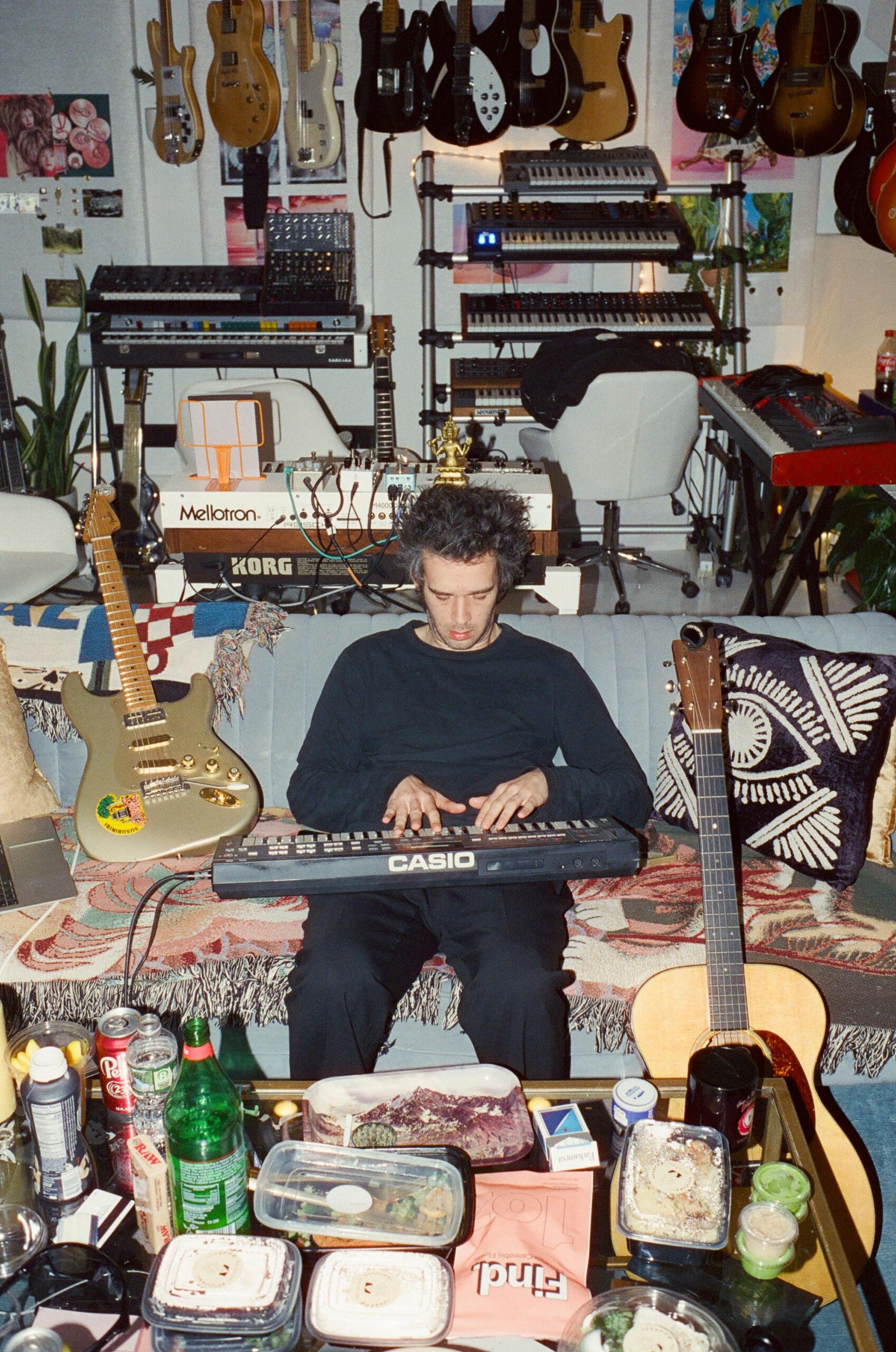 Matty Healy nga The 1975 luan në tastierë i rrethuar nga rrëmujë dhe instrumente muzikorë në atë që duket si një dhomë ndenjeje.