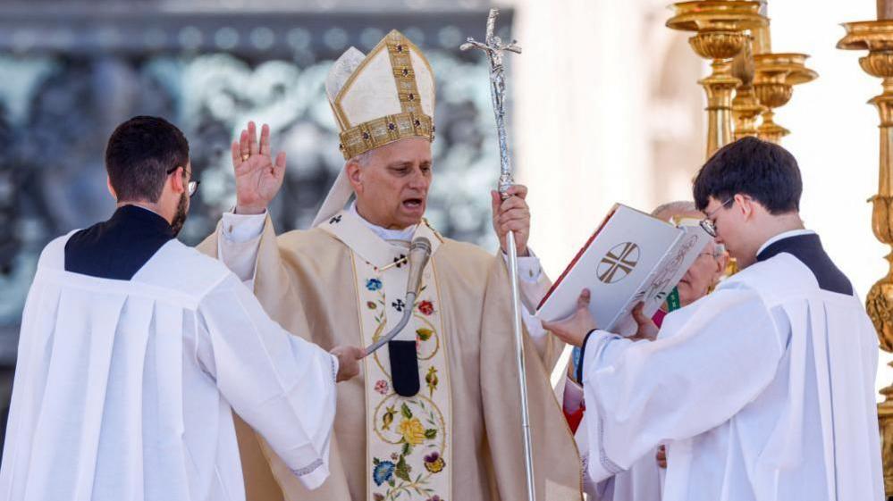 Papa Leo flet në mikrofon me njërën dorë të ngritur në një gjest bekimi