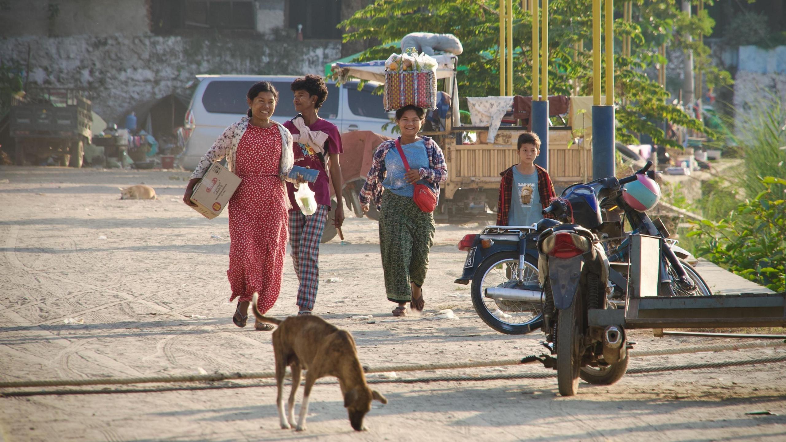 Njerëz që ecin në rrugë në Mandalay, pranë dy rrotave të parkuara. Në foto është edhe një qen që nuhat tokën.