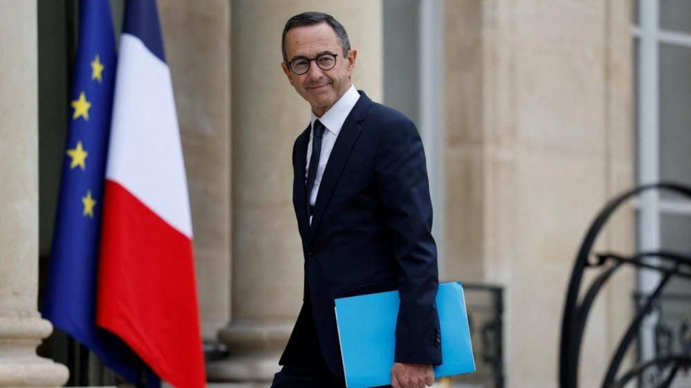 Bruno Retailleau, Ministri i Brendshëm Francez në largim, është fotografuar jashtë Pallatit Elysee përpara flamujve francezë dhe të BE-së. Ai është një burrë me flokë të errët me një kostum të errët, me syze dhe një dosje blu në dorë.