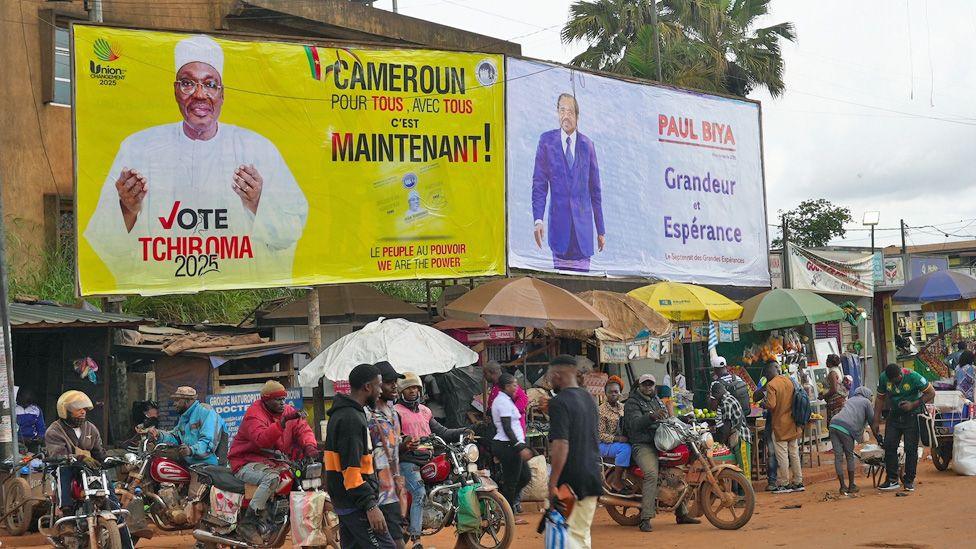 Njerëzit enden me motoçikleta në një rrugë me pluhur ku janë ngritur dy postera fushate - njëri për Biya-n dhe tjetri për Tchiroma-n.