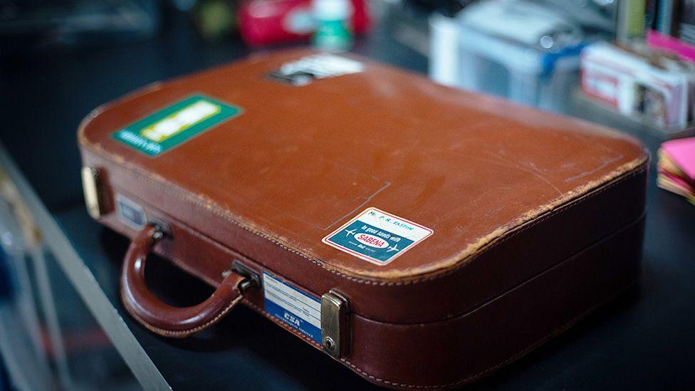 Një valixhe lëkure ngjyrë kafe e konsumuar me afishe udhëtimi, duke përfshirë 'Fly the Atlantic' dhe 'EAA', të vendosura mbi një sipërfaqe të errët. Sfondi është një ambient i brendshëm me rafte që mbajnë objekte të ndryshme të paqarta, përfshirë një shishe të kuqe.