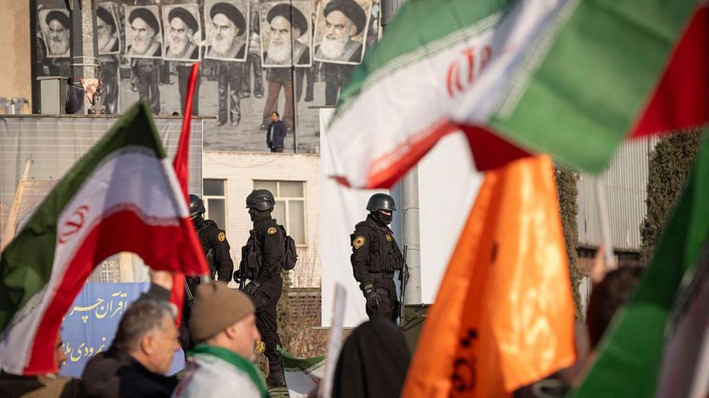 Oficerë policie të veshur të zinj me armë duke patrulluar një turmë që mbante flamuj iranianë.