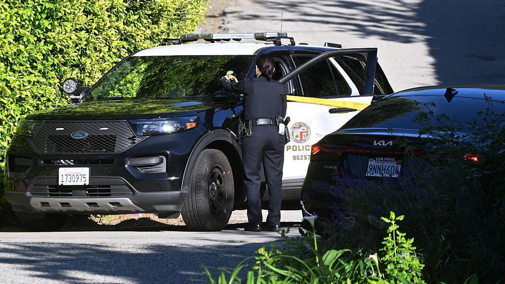 Një makinë policie është parkuar në një rrugë me një oficer që qëndron pranë saj