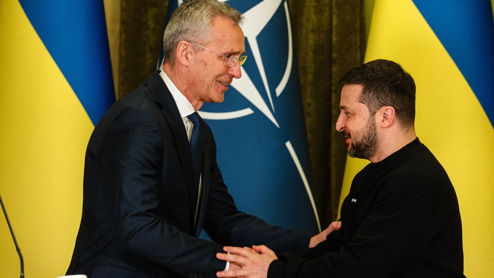 Ish-sekretari i përgjithshëm i NATO-s, Jens Stoltenberg, buzëqesh ndërsa shtrëngon duart me Presidentin ukrainas Volodymyr Zelensky përpara flamujve të NATO-s dhe Ukrainës në një takim në Kiev në maj 2023.