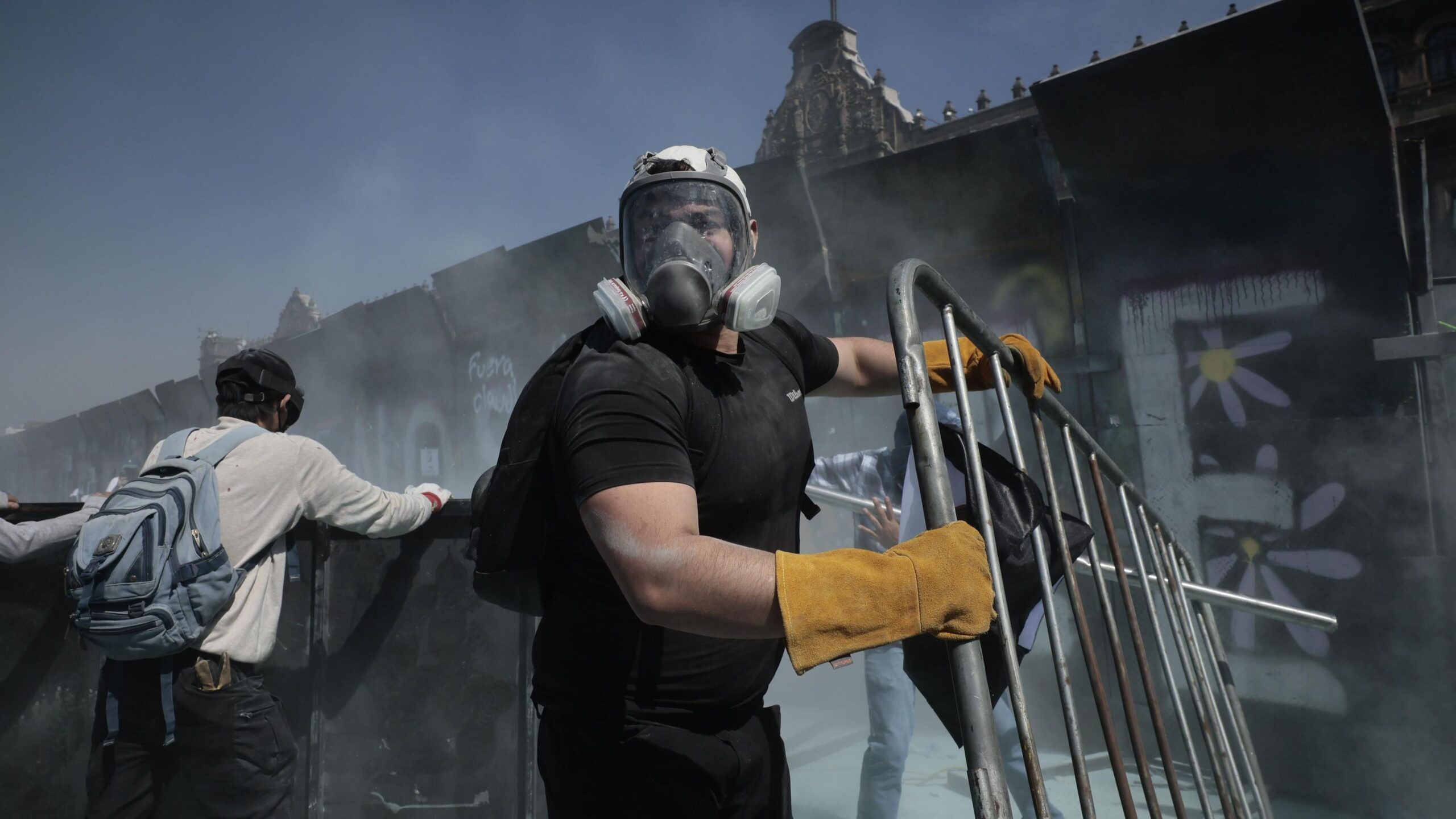Një protestues i veshur me maskë gazi dhe doreza ngjyrë mustardë mban lart një gardh gjatë një demonstrate pranë Pallatit Kombëtar në Meksikë.