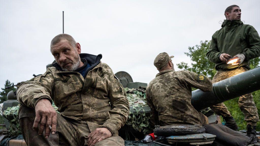 Ushtarë ukrainas të veshur me veshje të plota luftimi ulen mbi një top në një fushë me baltë.  