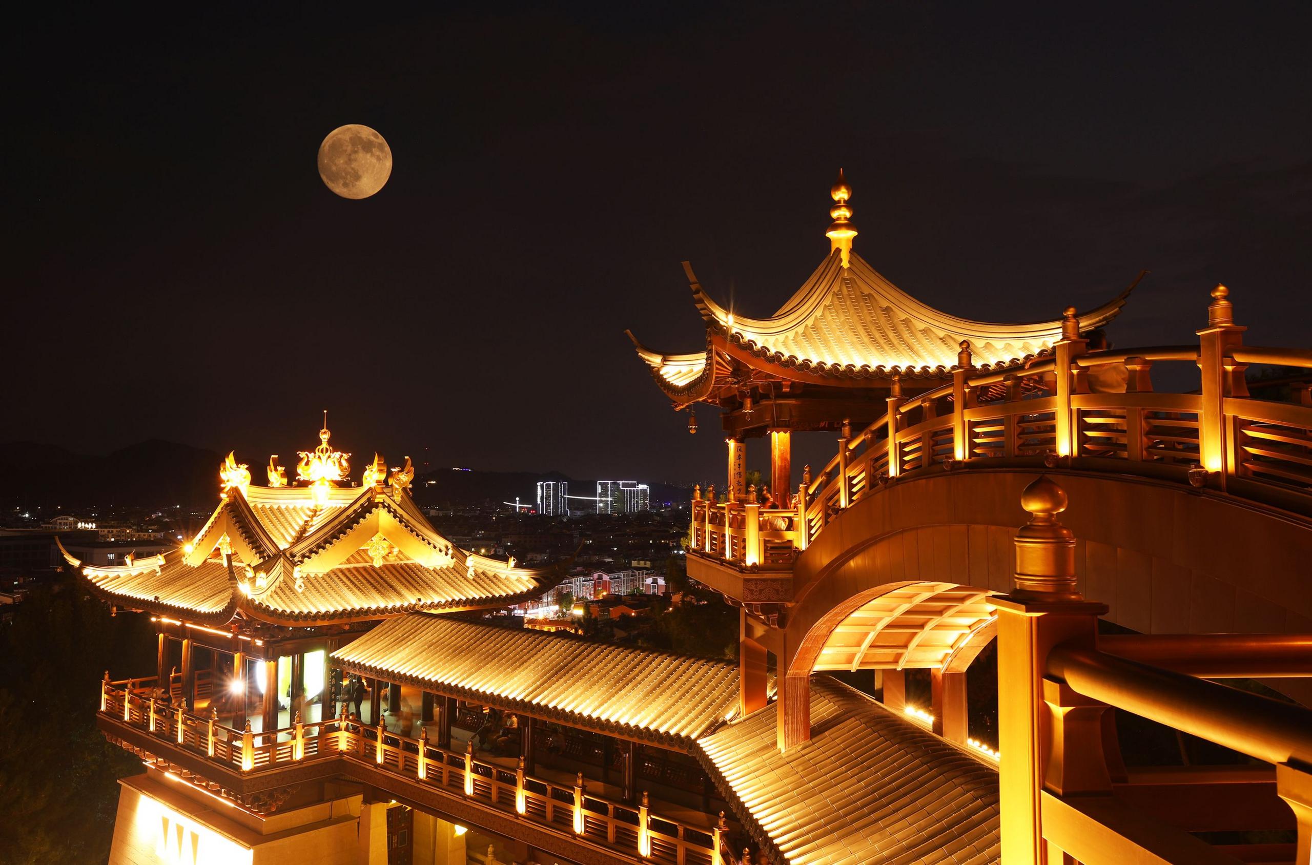 Superhëna e plotë në sfondin e një qielli të errët nate, duke fluturuar mbi një pavijon të ndriçuar mirë dhe të projektuar me zbukurime në Jinhua, Provinca Zhejiang.