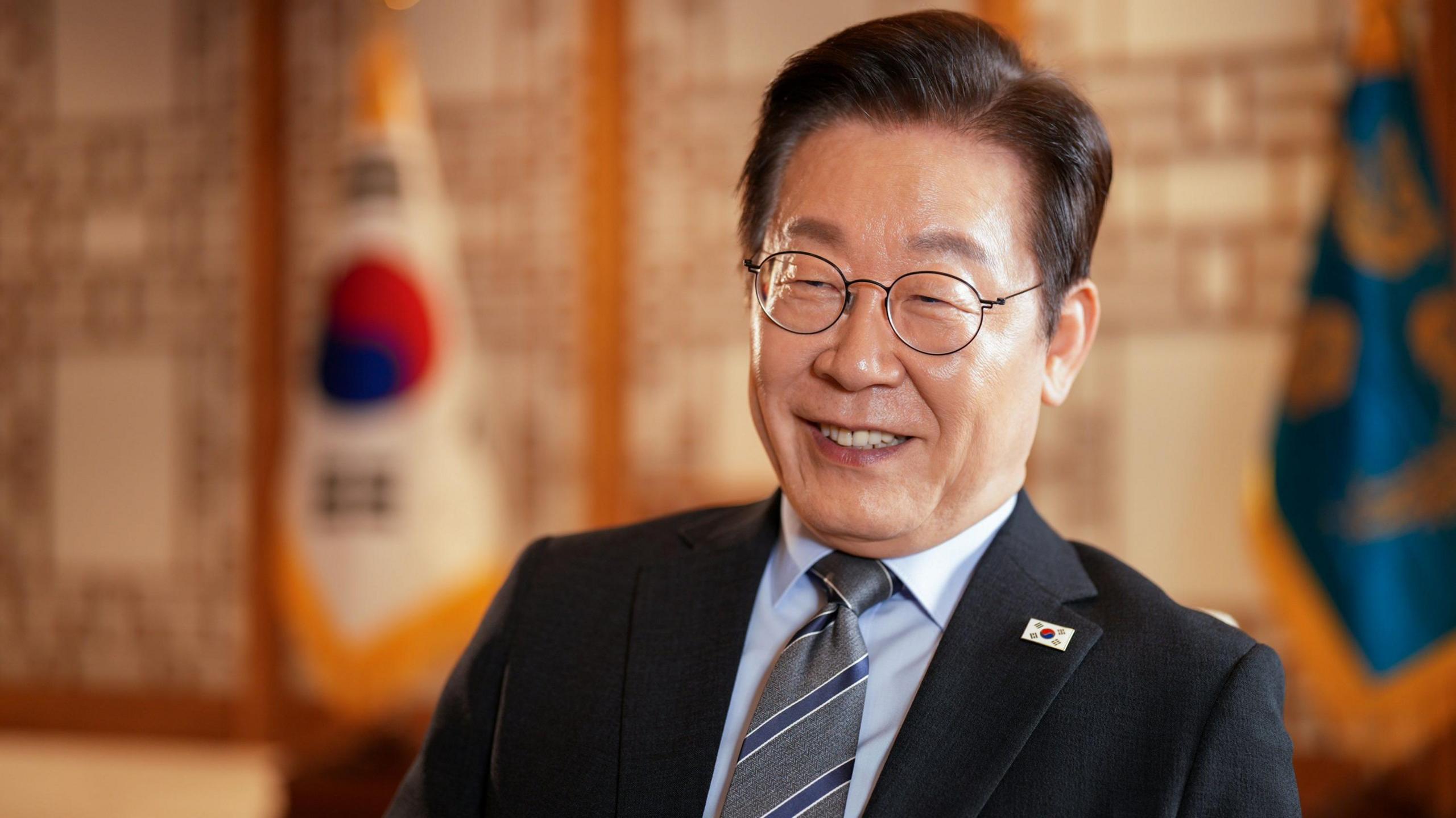 Presidenti Lee Jae Myung është ulur në një karrige i veshur me kostum të zi në zyrën e tij në Seul. Pas tij janë dy flamuj.