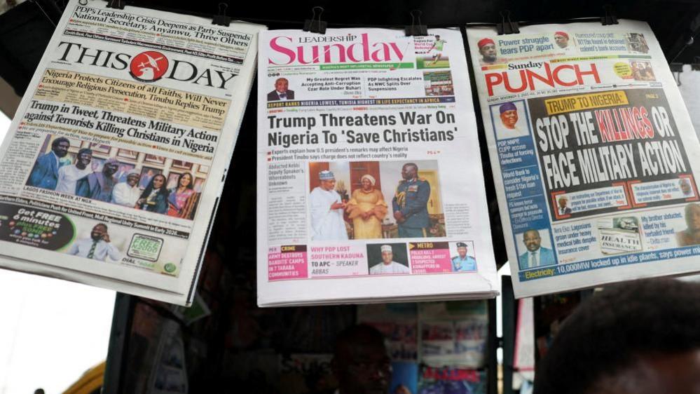 Gazetat me artikuj që raportojnë mesazhin e Presidentit të SHBA-së Donald Trump drejtuar Nigerisë në lidhje me trajtimin e të krishterëve varen në një stendë gazetash. Titujt kryesorë lexojnë: Trump kërcënon Nigerinë me luftë për të shpëtuar të krishterët; Ndaloni vrasjet ose përballuni me veprime ushtarake, Trump në Twitter kërcënon me veprime ushtarake.