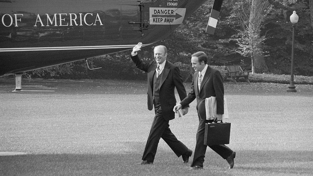 Presidenti Gerald Ford përshëndet me dorë ndërsa kalon pranë helikopterit presidencial me Dick Cheney-n që ecën pranë tij, treguar në vitin 1975 në një foto bardhë e zi.