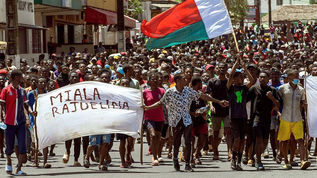Dhjetëra protestues mund të shihen duke marshuar përgjatë një rruge. Në pjesën e përparme janë njerëz që mbajnë një banderolë ku shkruhet "Rajoelina jep dorëheqjen" në gjuhën malagaziane. Mund të shihet edhe një flamur i Madagaskarit.