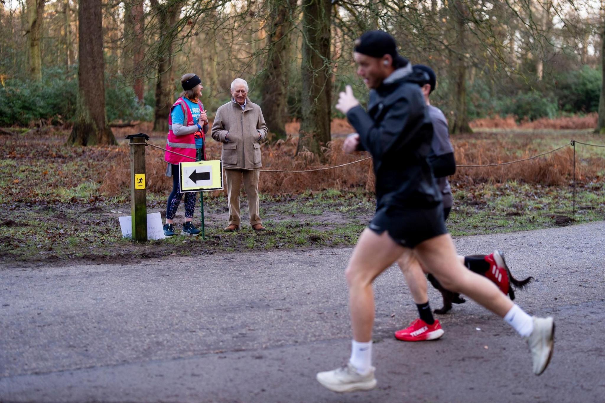 Dy vrapues dhe një qen në një rrugë të shtruar me metal në Sandringham Parkrun. Mbreti Charles dhe një vullnetar i garës po qëndrojnë në buzë të rrugës, me një pyll pas tyre.