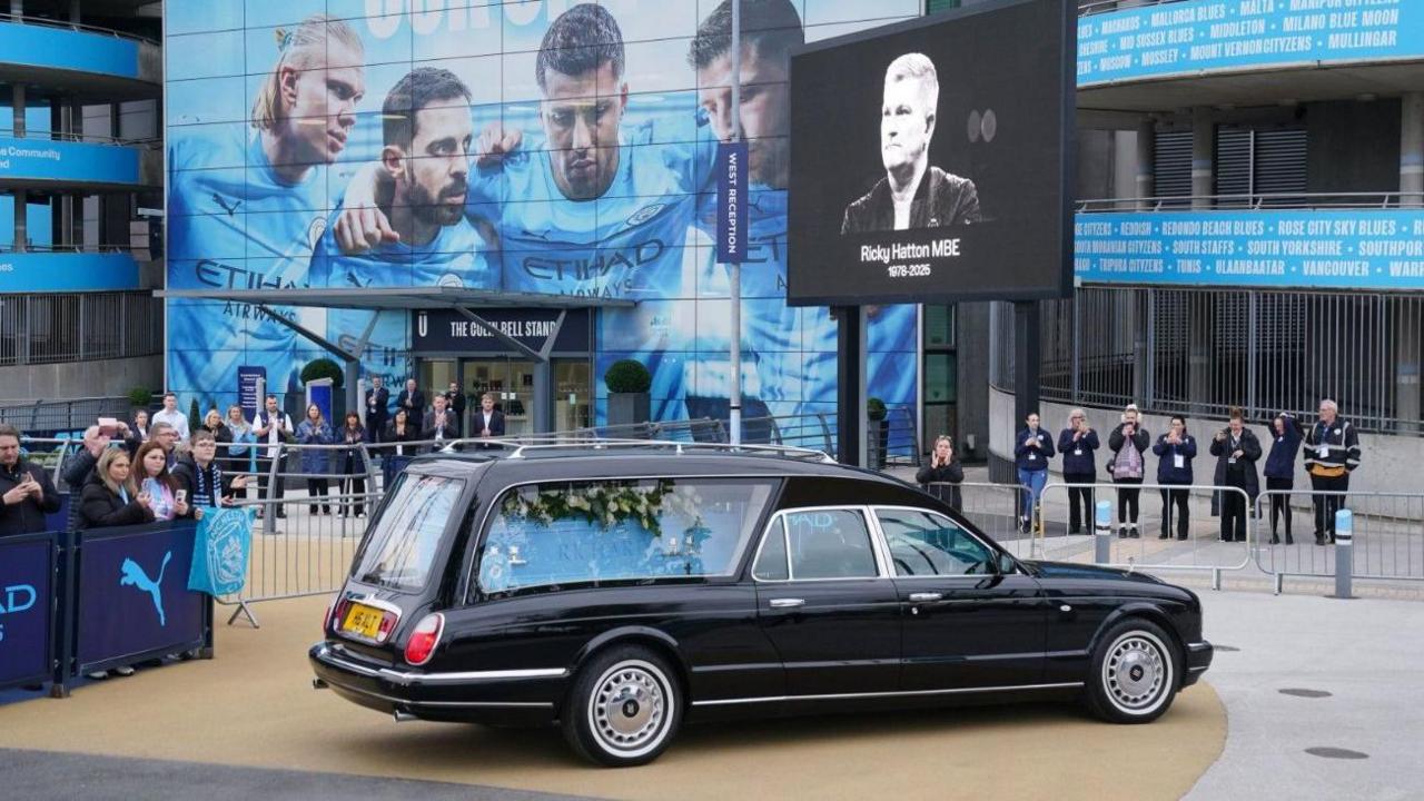 Karroca e zezë është parkuar jashtë stadiumit Etihad. Një fotografi bardhë e zi e Hattonit është në ekranin përpara stadiumit.