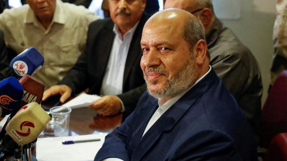 Një fotografi e Khalil al-Hayya-s. Ai është ulur në një konferencë për shtyp dhe tre burra të tjerë mund të shihen në sfond pas tij. Ai ka veshur një kostum blu të errët.