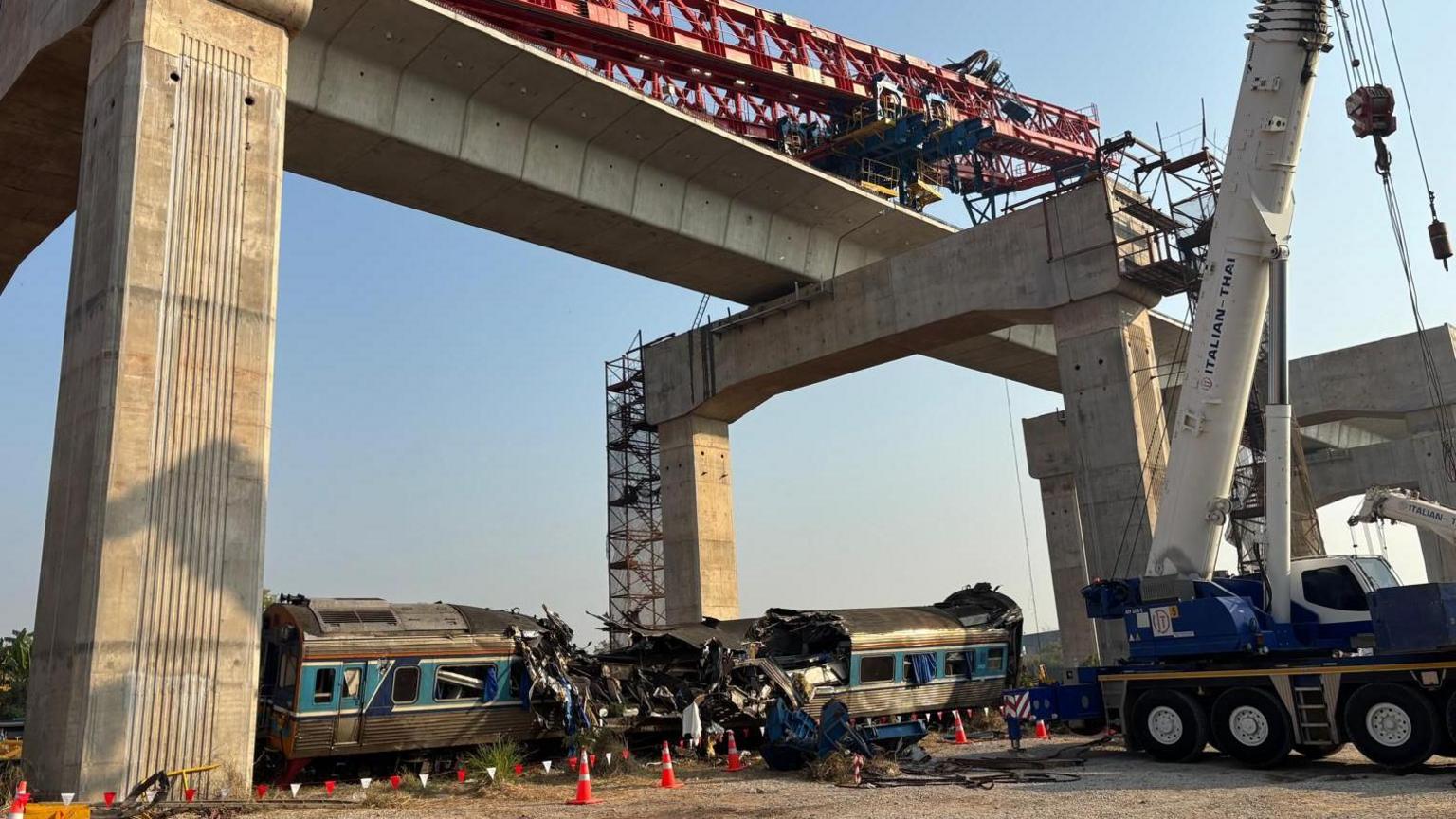 Një vinç i shtrirë anash përgjatë binarëve hekurudhorë sipër dhe një tren blu i rrëzuar në tokë poshtë. Disa nga përplasjet e trenave janë shkatërruar plotësisht.