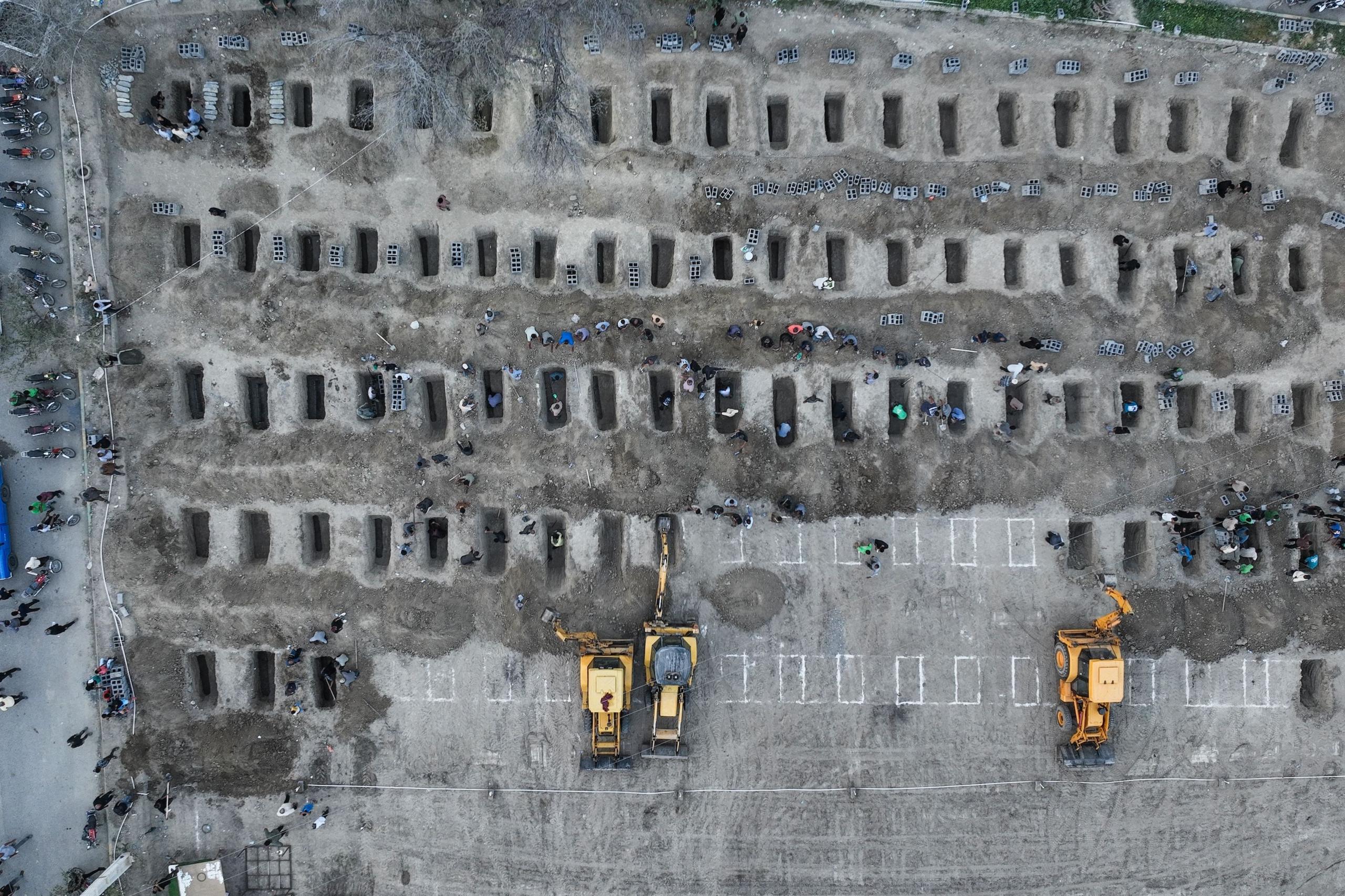 Një fotografi ajrore tregon një sërë varresh të gërmuara në tokë me njerëz përreth tyre. Traktorë të verdhë janë gjithashtu në pamje.
