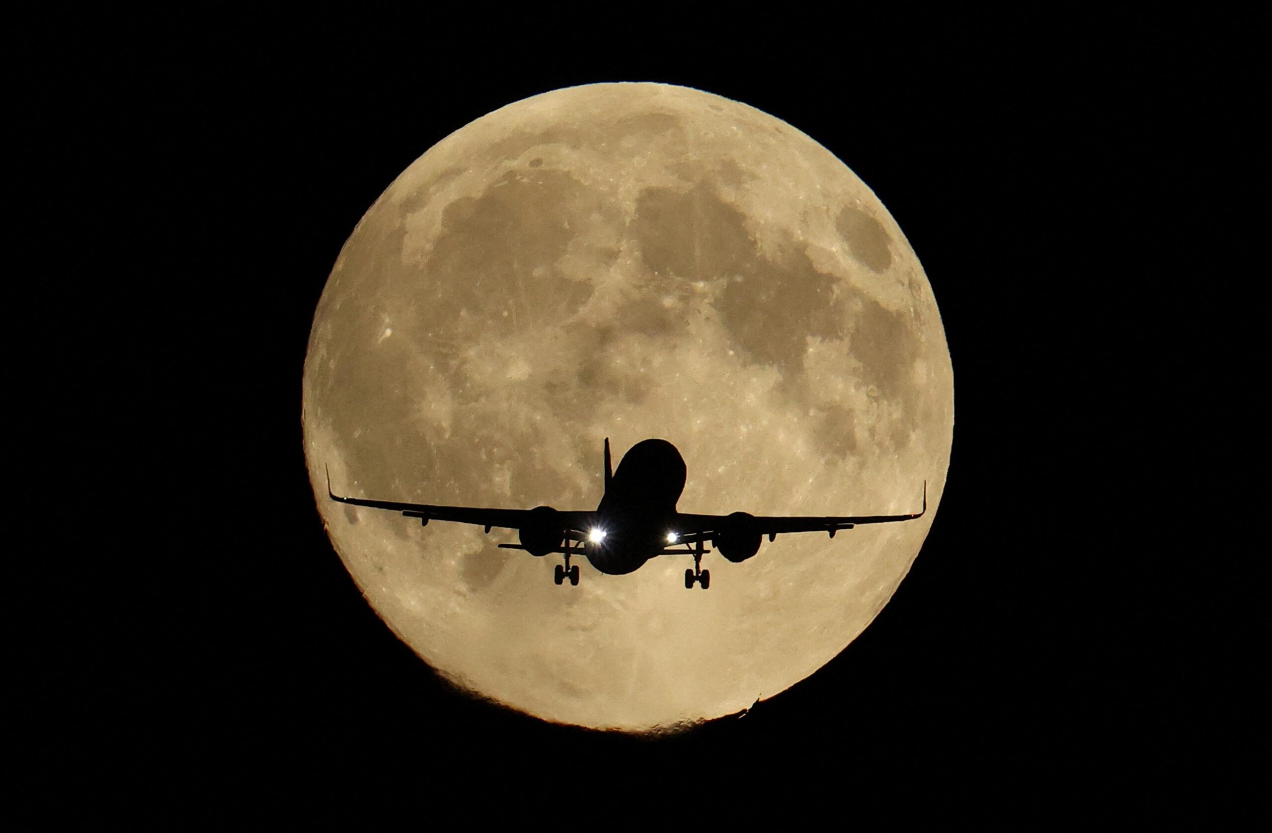 Një aeroplan pasagjerësh shfaqet në siluetë përpara një hëne të plotë në qiellin e zi të natës