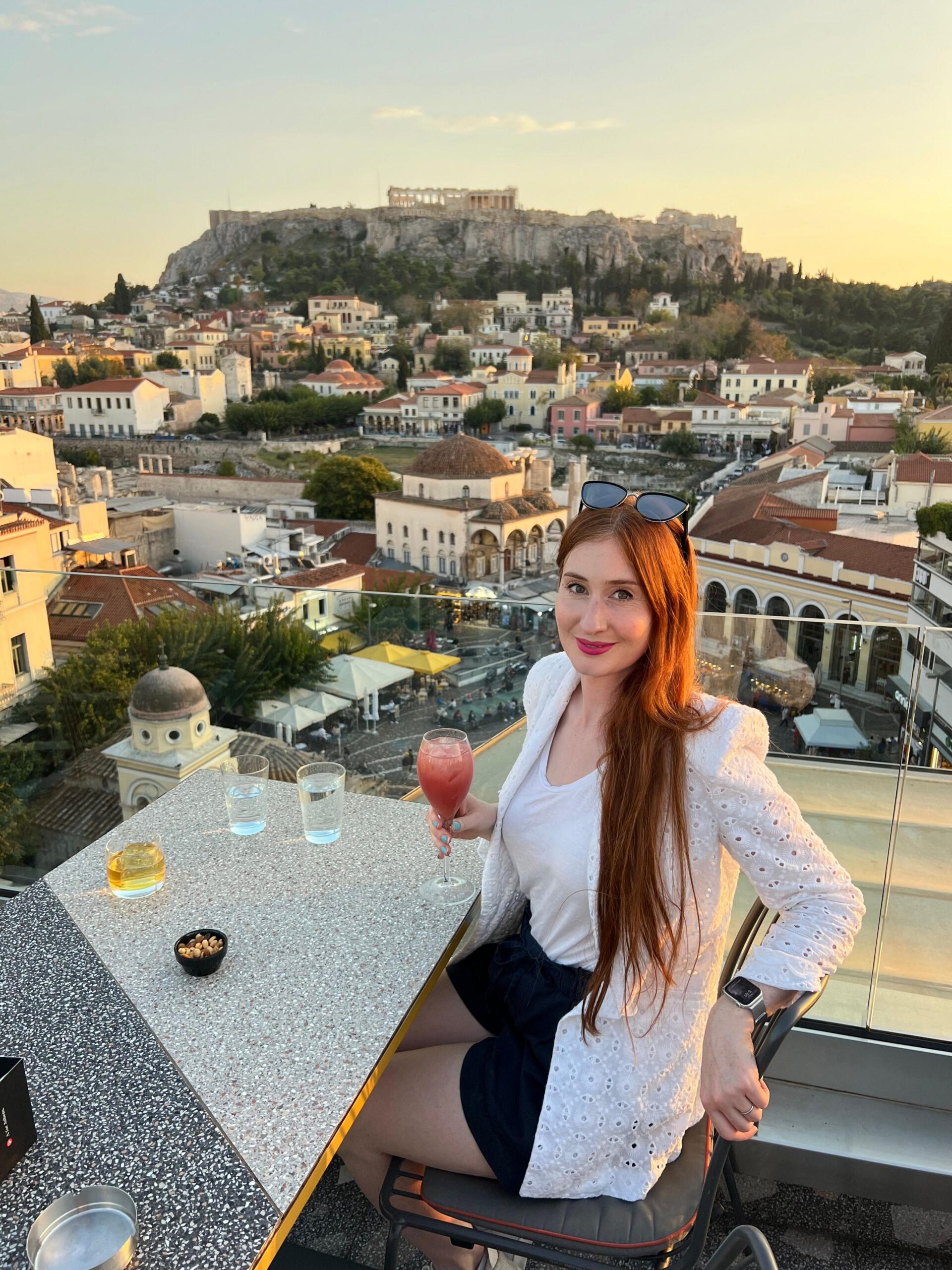 Amber, një grua e re me flokë të kuq, e ulur në një tavolinë ngrënieje jashtë në Athinë. Akropoli është i dukshëm në sfond.