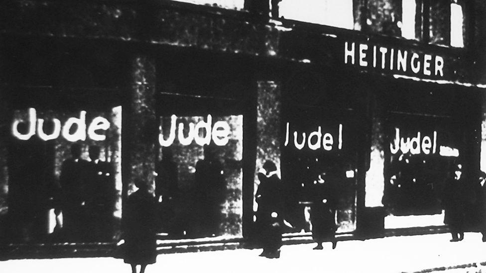 Fotografi bardhë e zi e një vitrine dyqani me etiketën "HEITINGER". Dritaret janë vandalizuar me fjalën "Jude" (gjermanisht për hebre) të shkruar me shkronja të mëdha të bardha. Disa njerëz shihen duke kaluar pranë dyqanit ose duke qëndruar pranë tij.
