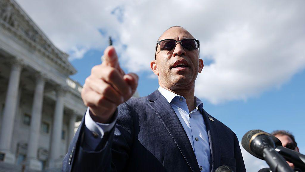 Udhëheqësi i pakicës në Dhomën e Përfaqësuesve të SHBA-së, Hakeem Jeffries, demokrat i Nju Jorkut, vesh një kostum blu të errët dhe syze dielli ndërsa qëndron në shkallët e Kapitolit të SHBA-së në Uashington. Ai po tregon me gishtin tregues të djathtë ndërsa flet me gazetarët.