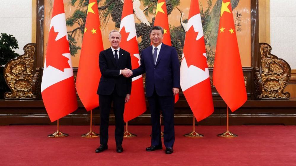 Kryeministri kanadez Mark Carney buzëqesh ndërsa shtrëngon duart me Presidentin e Kinës Xi Jinping në Sallën e Madhe të Popullit në Pekin. Përballë kameras, ata qëndrojnë në një qilim të kuq para flamujve kanadezë dhe kinezë.