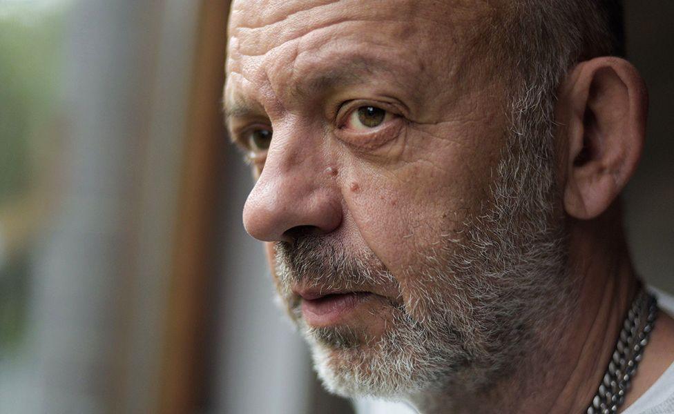 Pamje nga afër e Antony Easton, një burri tullac me mjekër gri, që shikon nga dritarja. Fytyra e tij është kthyer pak anash, duke nxjerrë në pah njollat e moshës dhe rrudhat. Ai mban veshur një këmishë të bardhë dhe një varëse argjendi. Sfondi është pak i turbullt, duke e tërhequr vëmendjen te tiparet e fytyrës së tij.