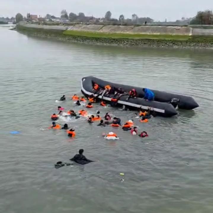 Njerëz, kryesisht të veshur me jelekë shpëtimi portokalli, notojnë drejt një gomone të zezë në një kanal.