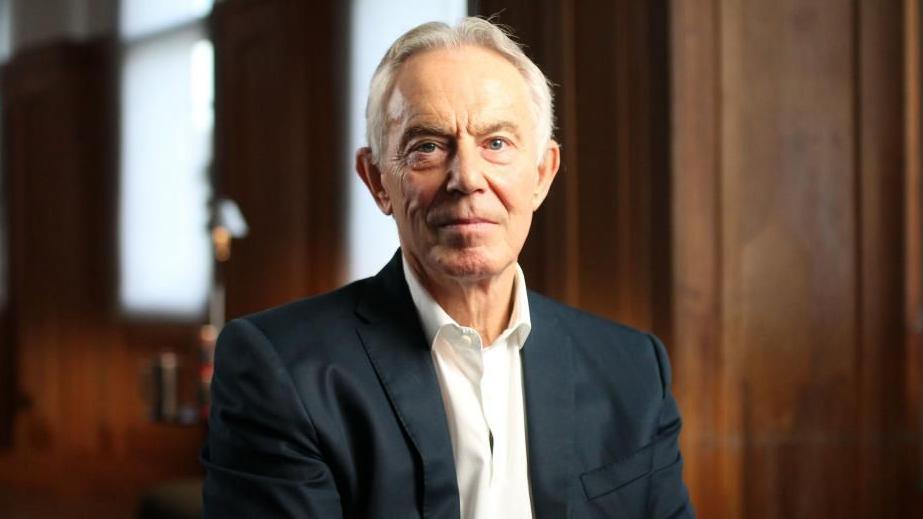 Një imazh i ish-kryeministrit të Mbretërisë së Bashkuar, Tony Blair, duke parë kamerën i veshur me kostum të zi dhe këmishë të bardhë në një dhomë me mure druri.