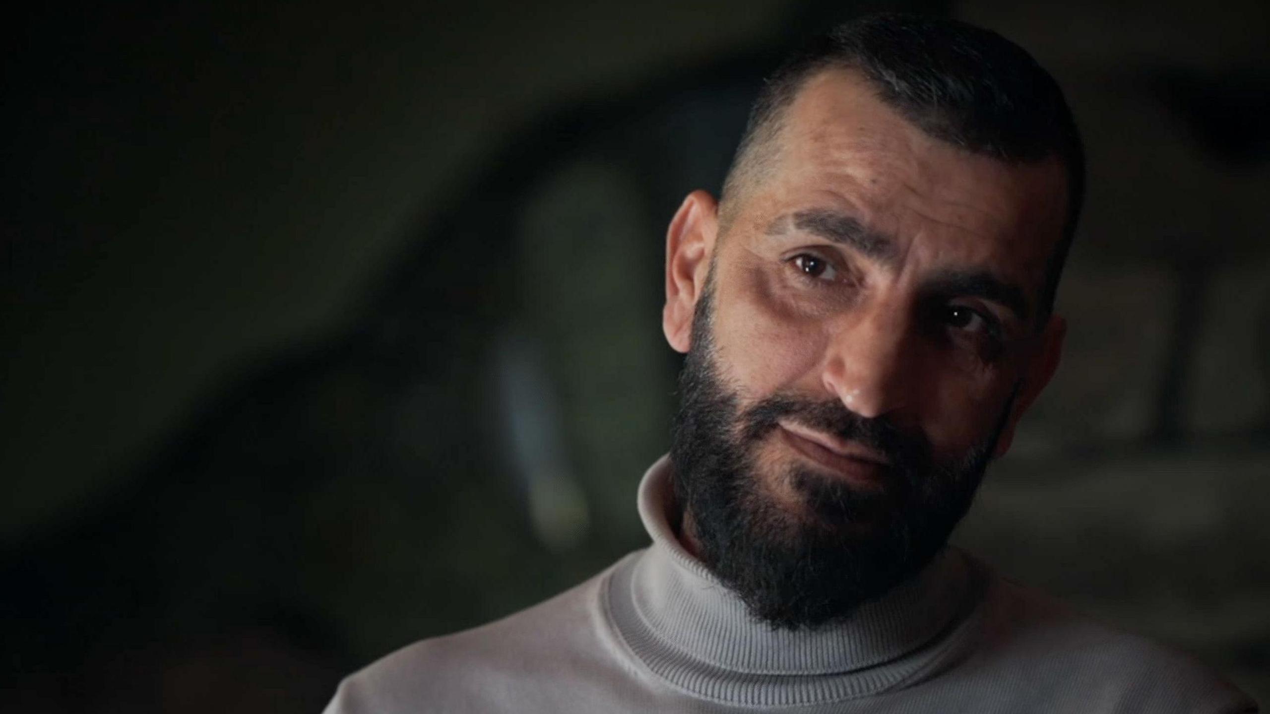 Habib me një bluzë të bardhë me jakë të rrumbullakët duke folur para kamerës në një sfond të errët. Ai ka mjekër dhe mustaqe të errëta.