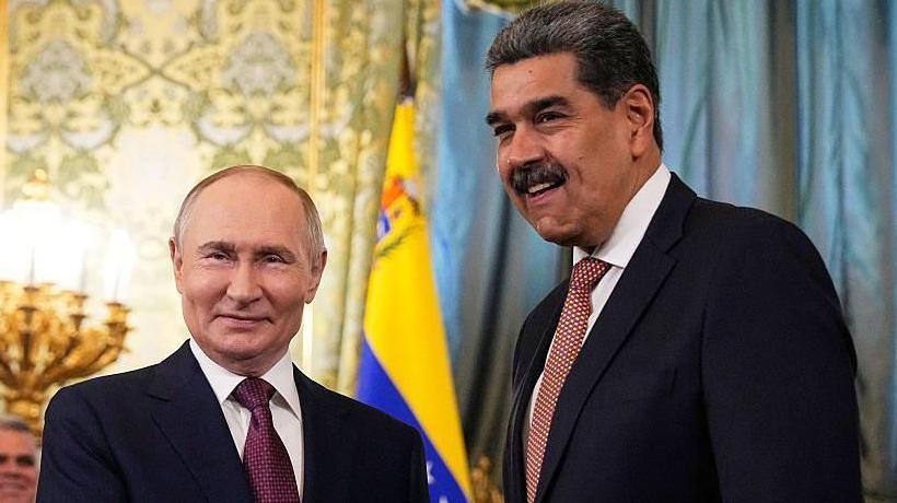 Presidenti rus Vladimir Putin, i veshur me një kostum të errët me kravatë ngjyrë burgundy me pika të bardha, shtrëngon duart me Presidentin e Venezuelës Nicolás Maduro, i cili vesh një kostum të errët dhe një kravatë portokalli me pika të bardha.