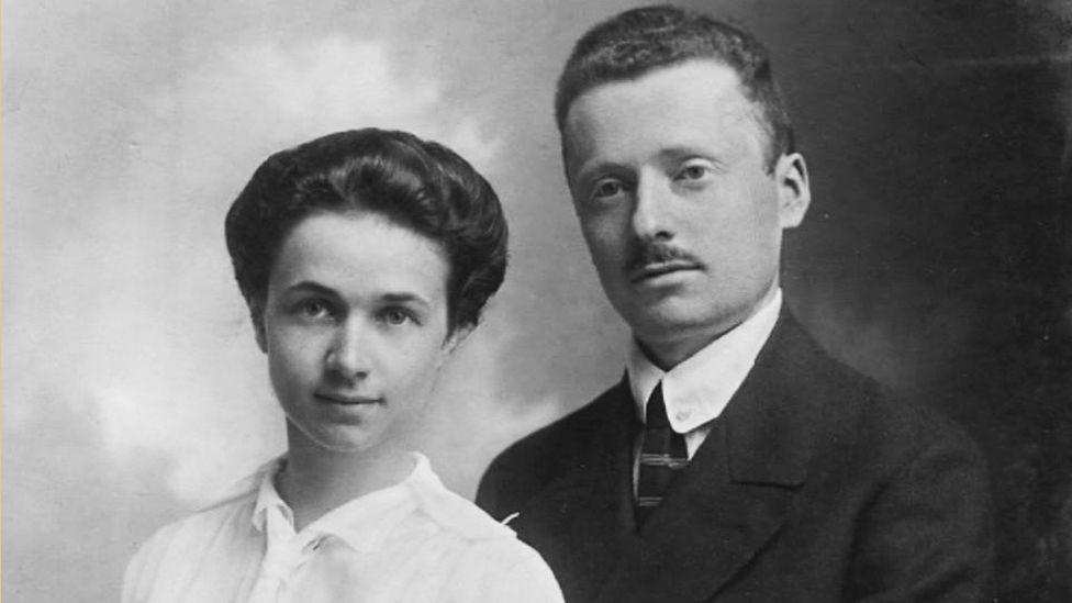 Fotografi bardhë e zi e Hildegarde dhe Rudolf Eisner, të cilët qëndrojnë pranë njëri-tjetrit. Hildegarde është në të majtë dhe mban veshur një bluzë të çelët me jakë të lartë, ndërsa Rudolf është veshur me një kostum të errët me këmishë dhe kravatë të bardhë. Sfondi është i thjeshtë me hije të lehta.